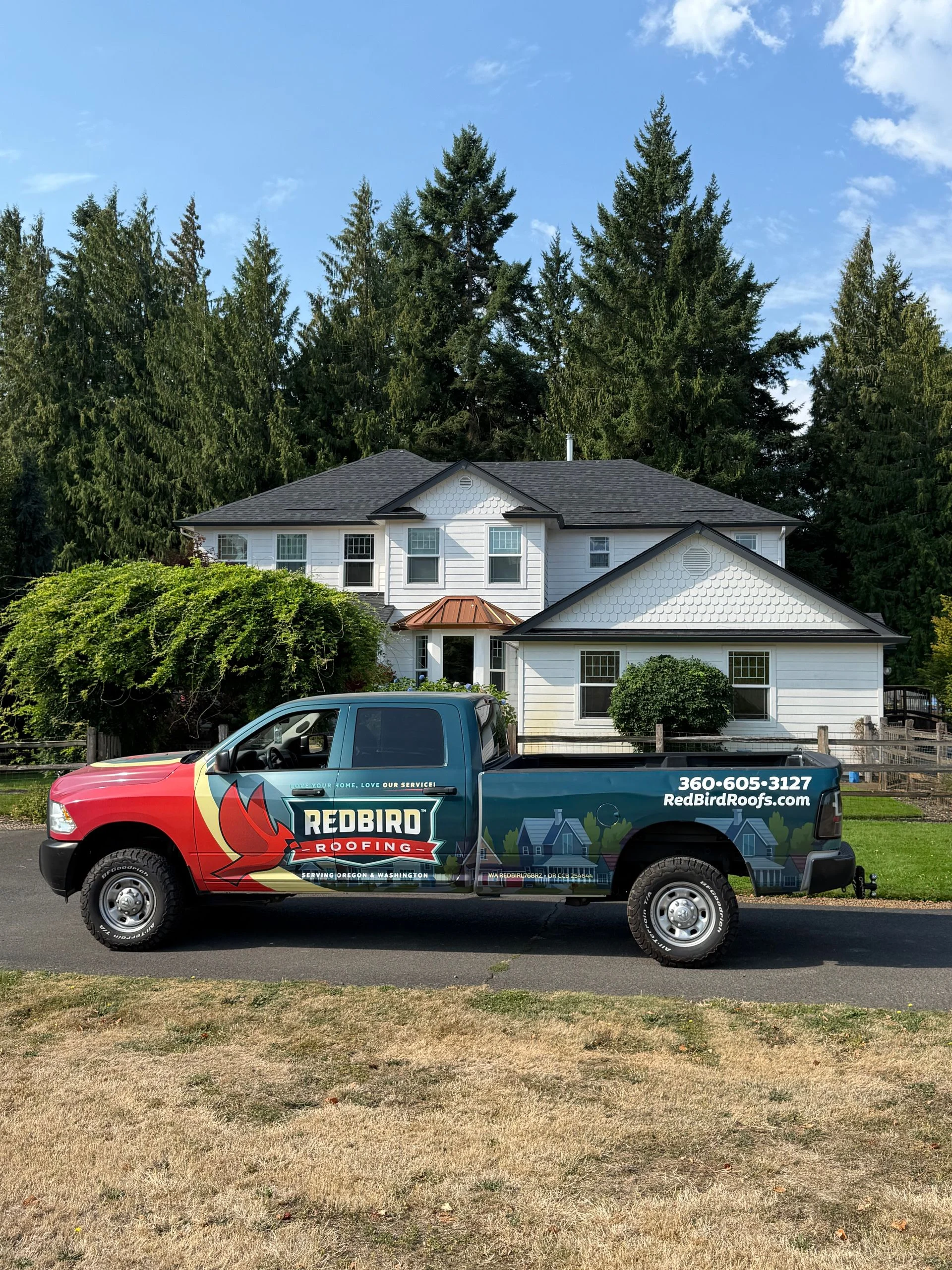 A Redbird Electric truck with a red and green wrap parked in front of a white two-story house surrounded by tall trees.