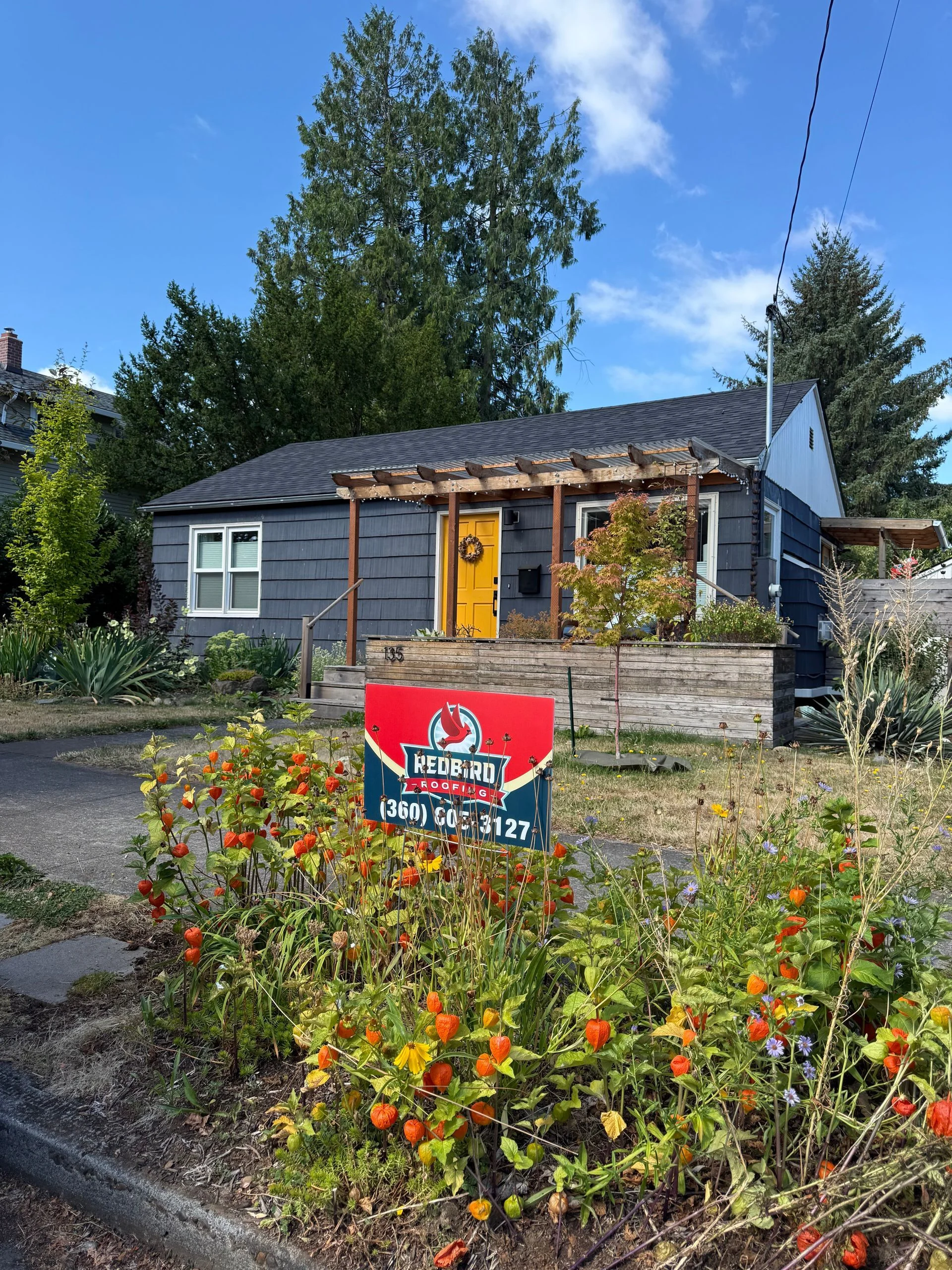 A grey house with a bright yellow front door, a wooden pergola, and a sign in the foreground amidst a garden.