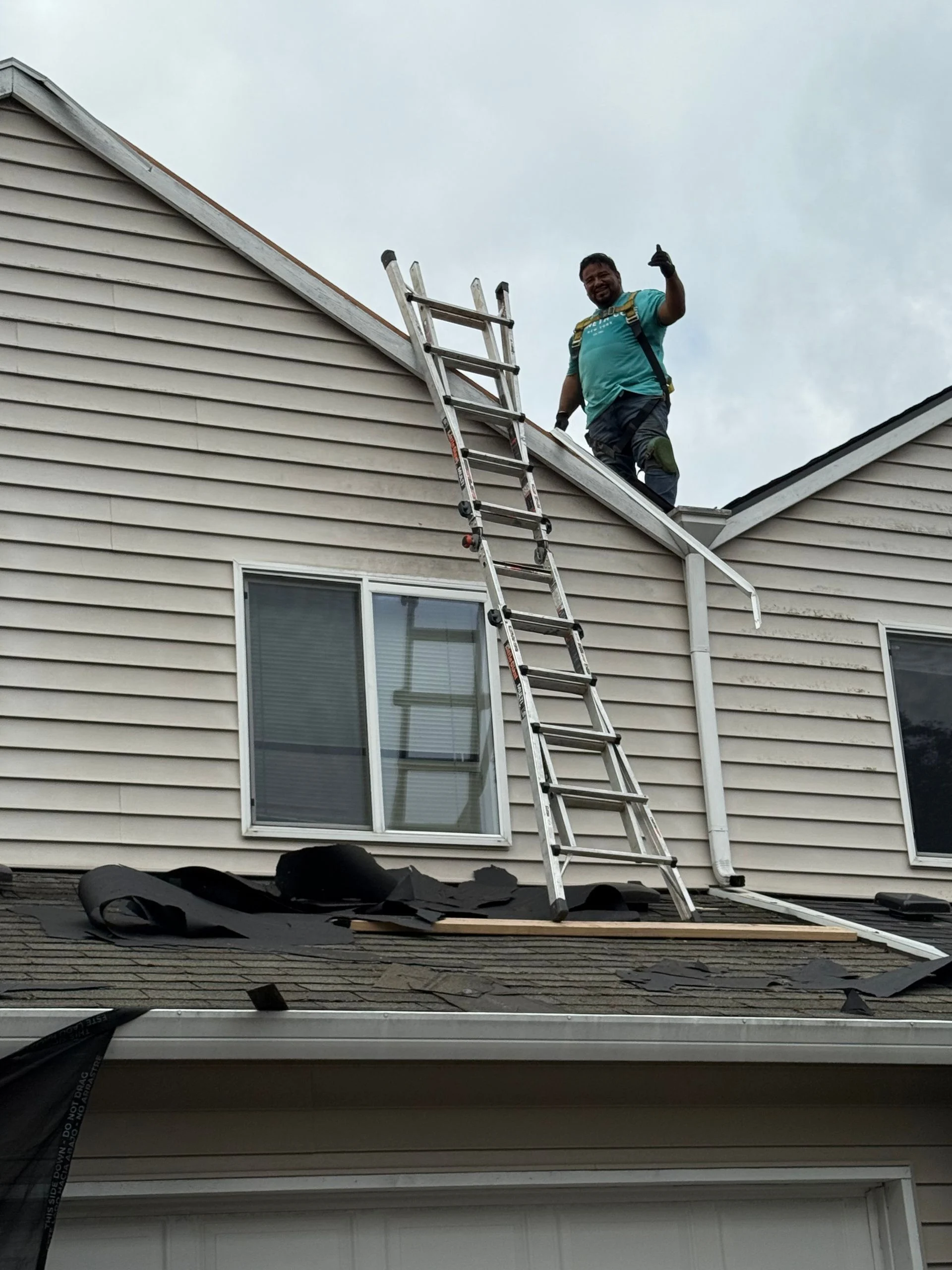 A worker stands on a roof next to an extended ladder, giving a thumbs-up gesture against a residential siding exterior.