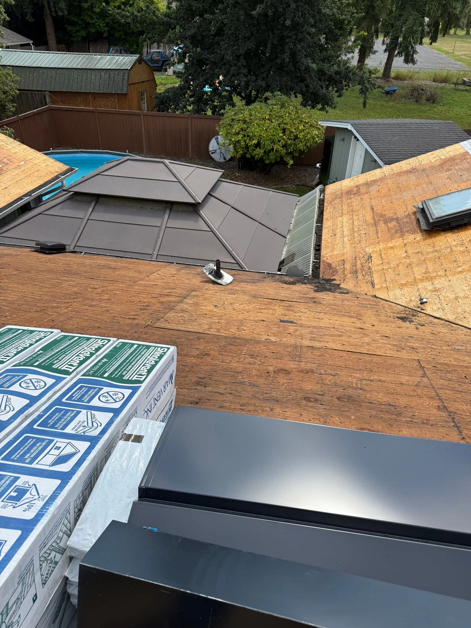 A high-angle view of a residential roof under construction, featuring exposed plywood sheathing and stacked shingles.