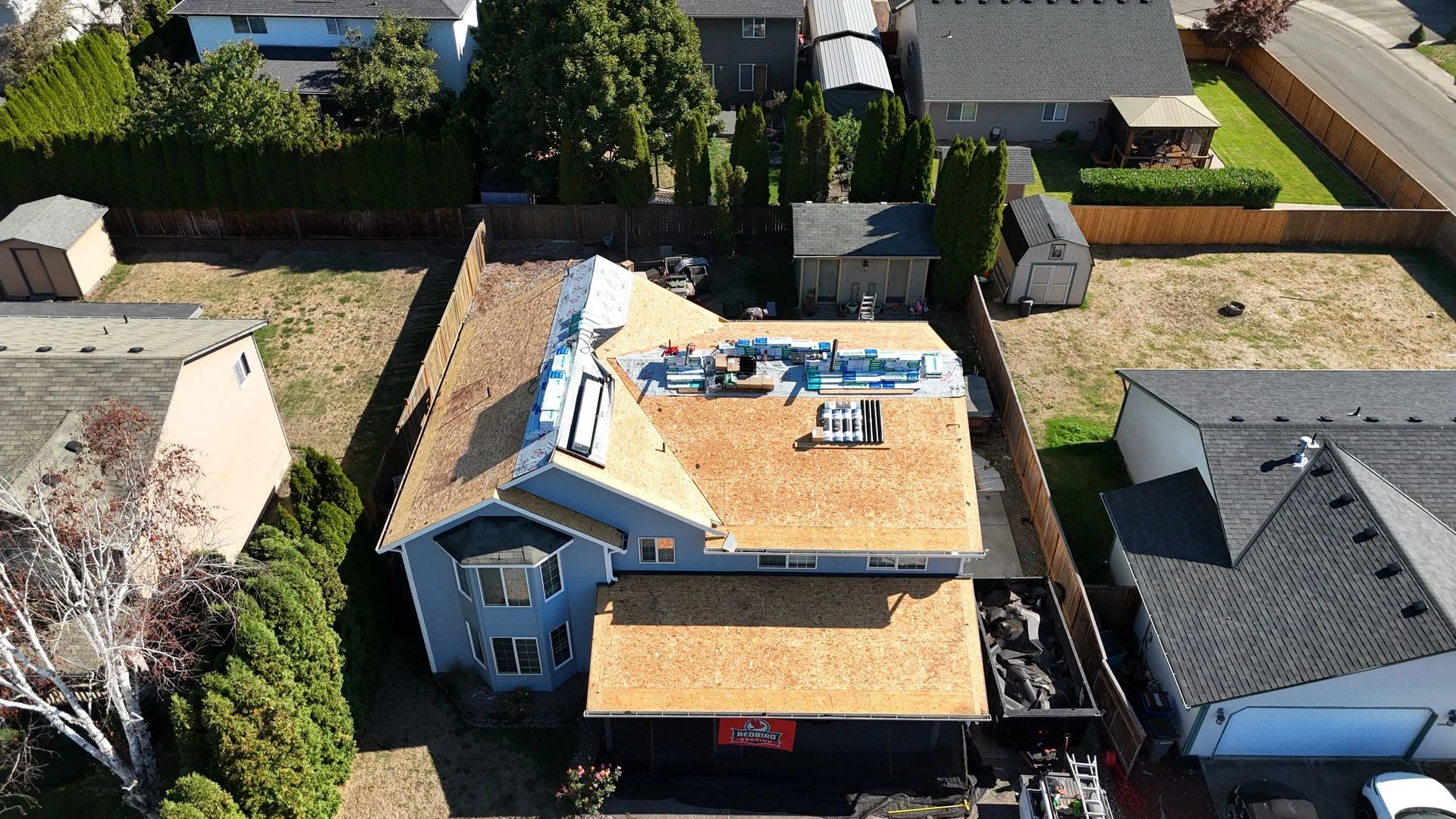 An aerial view of a house undergoing roof replacement, featuring exposed wood decking, construction materials, and a dumpster.