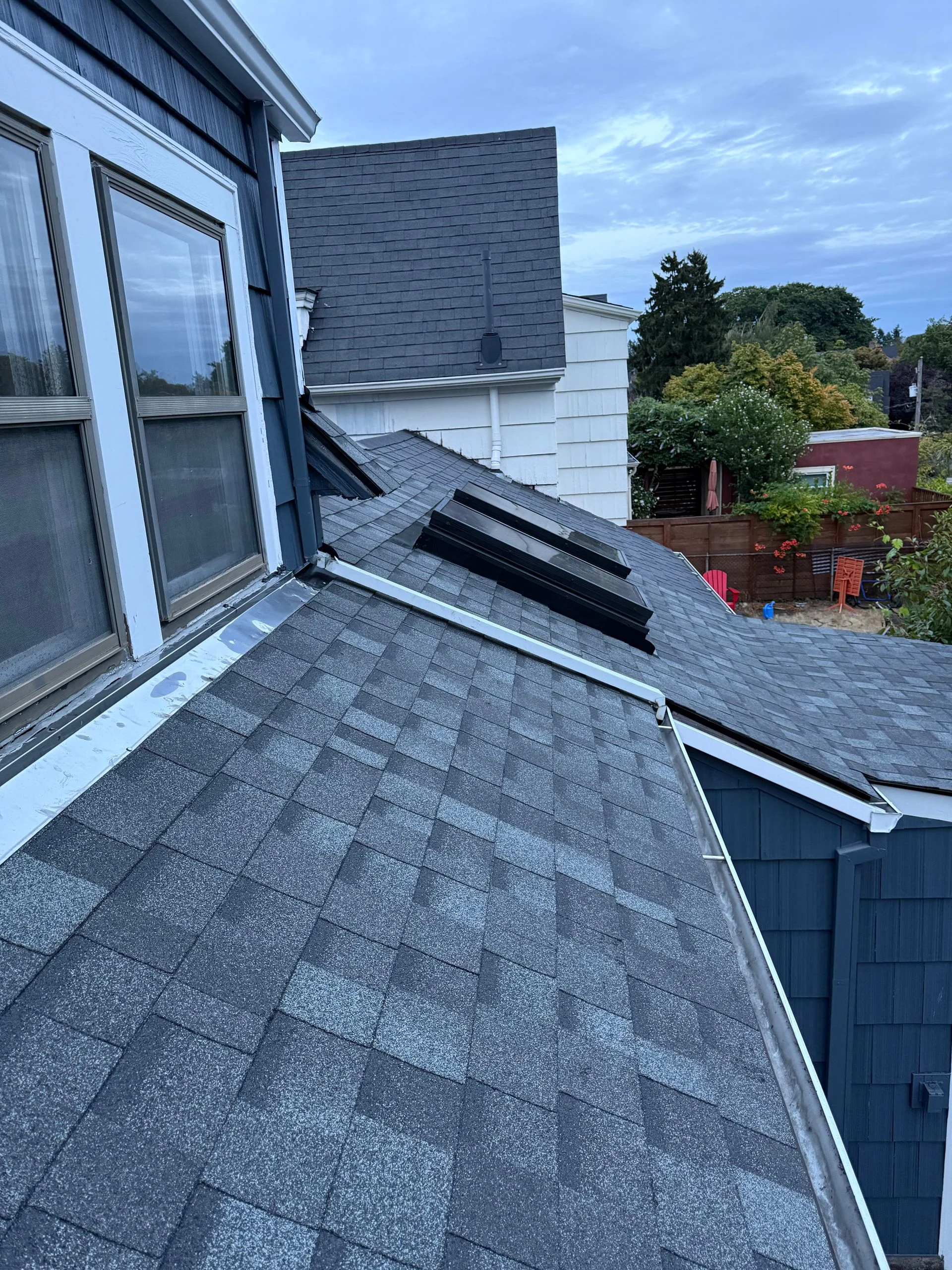 A high-angle view of a gray shingled residential roof with skylights, showing a dormer window and adjacent building.