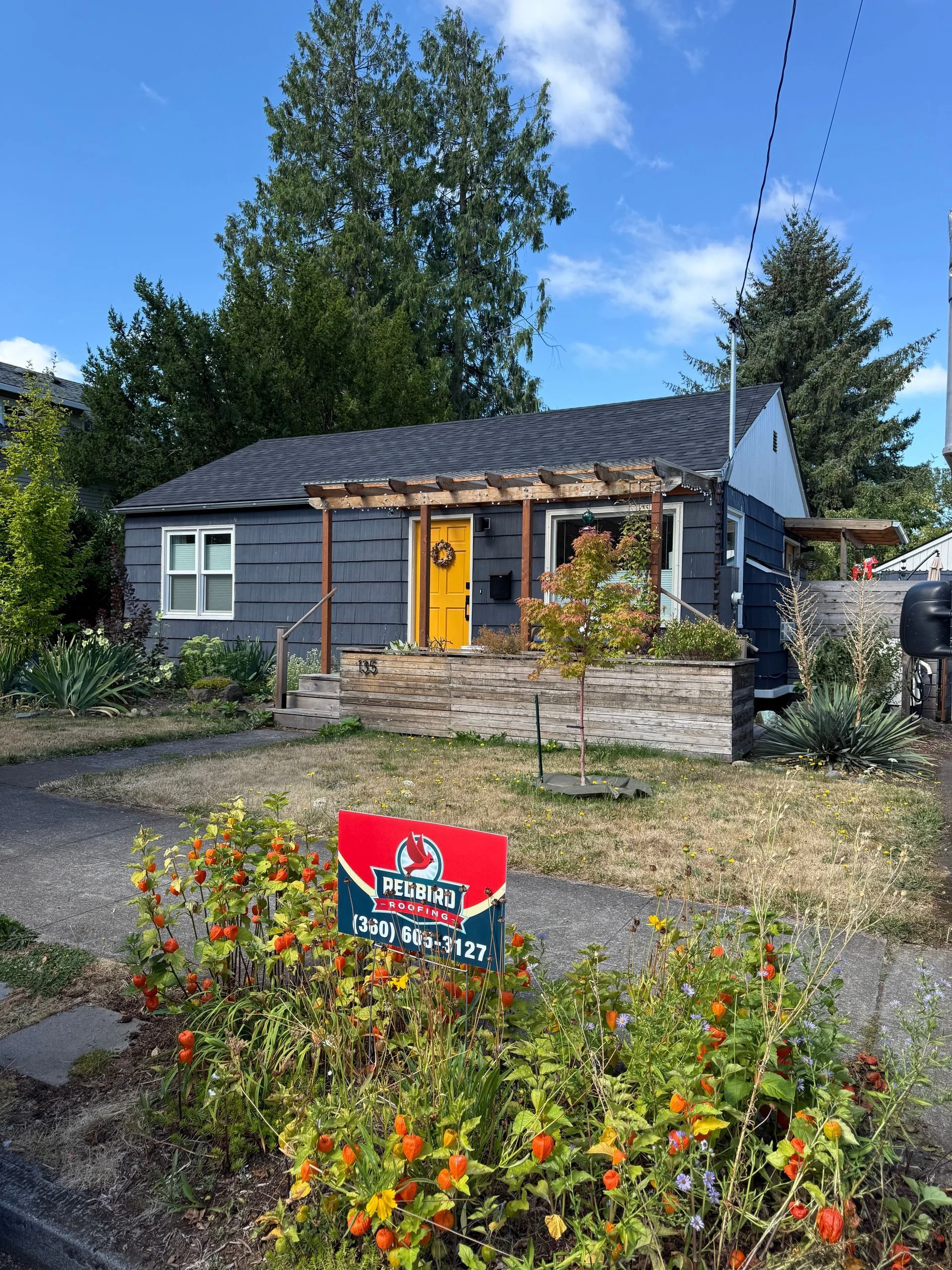 A single-story dark grey house with a yellow front door, a wooden pergola, and a 