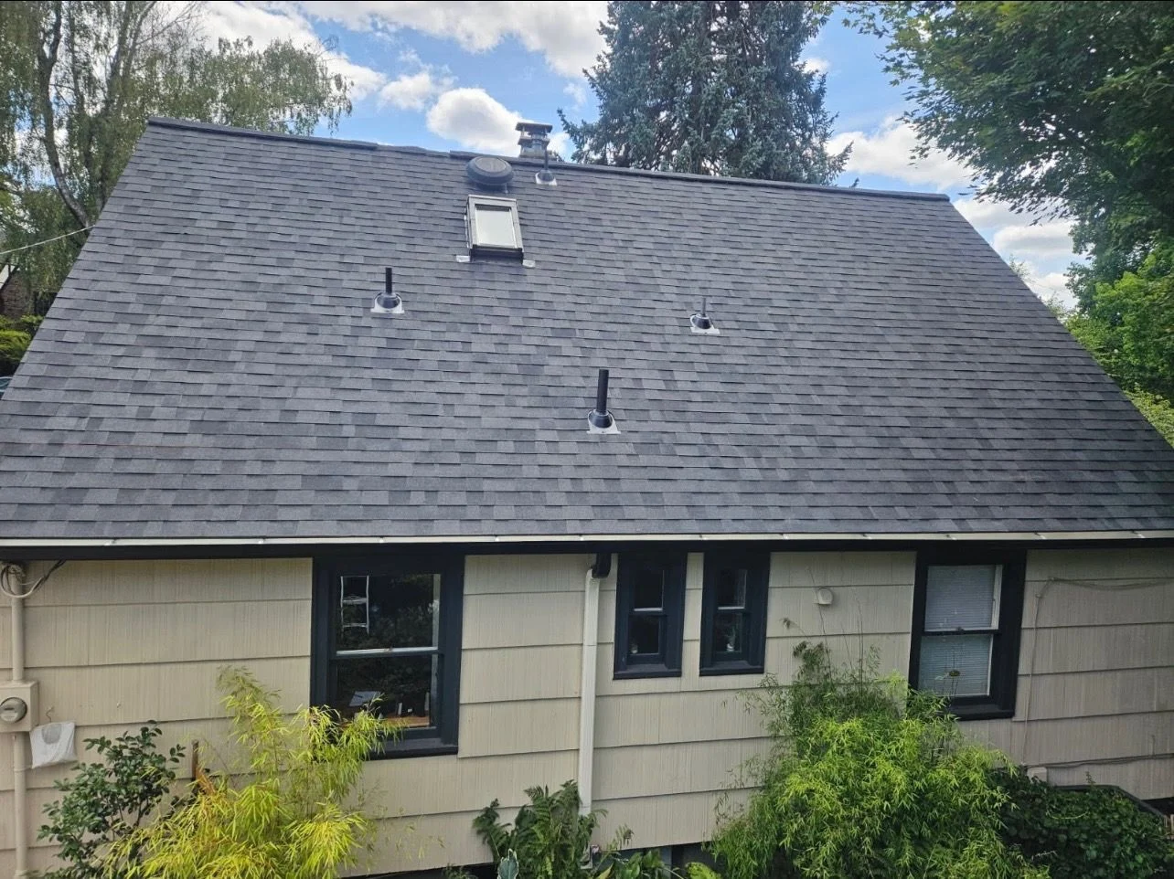 A rear view of a house with light-colored siding, dark-trimmed windows, and a dark shingled roof with several vents.