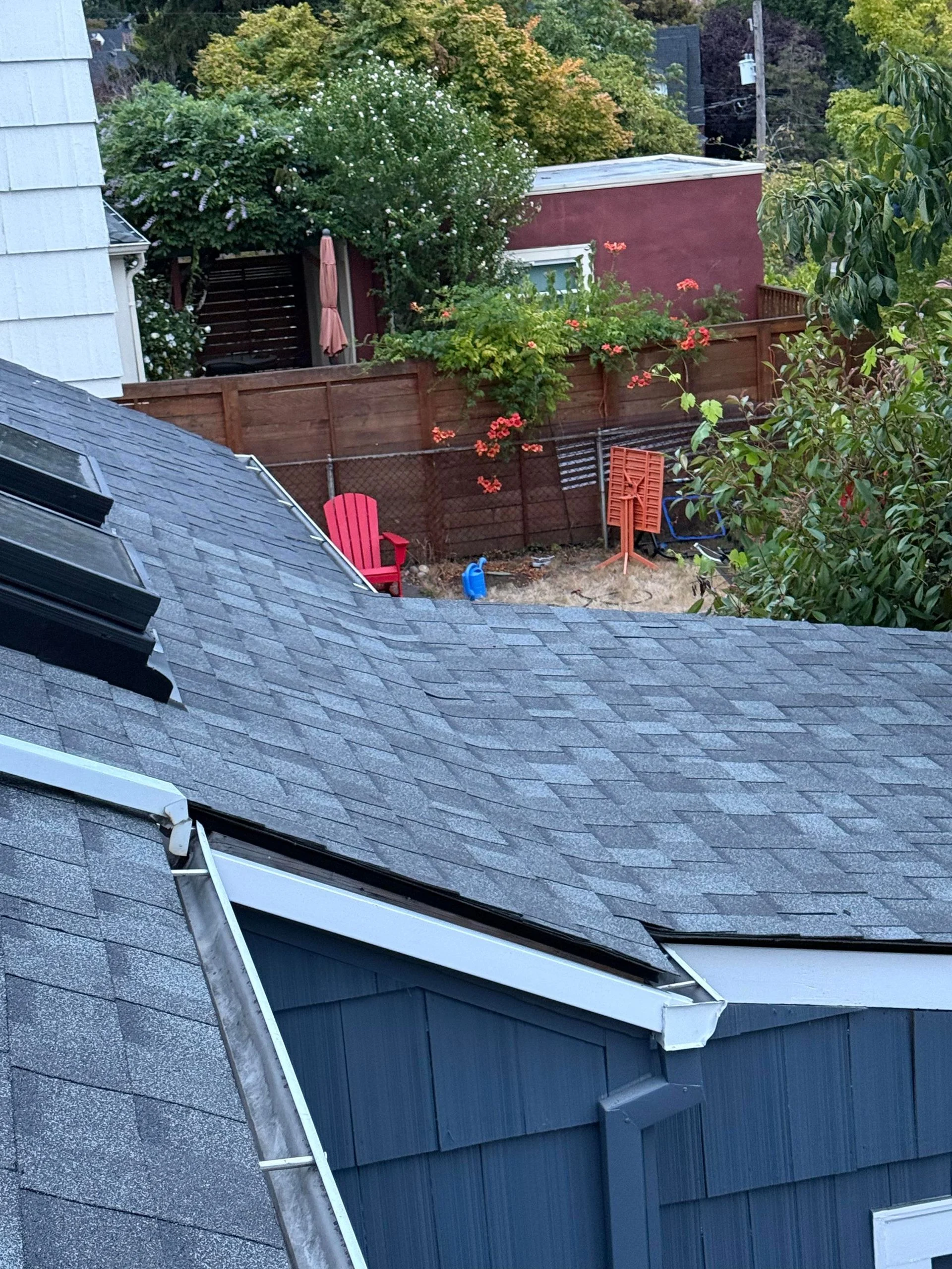 An elevated view of a gray shingled roof with a skylight, overlooking a backyard with a red chair and a wooden fence.