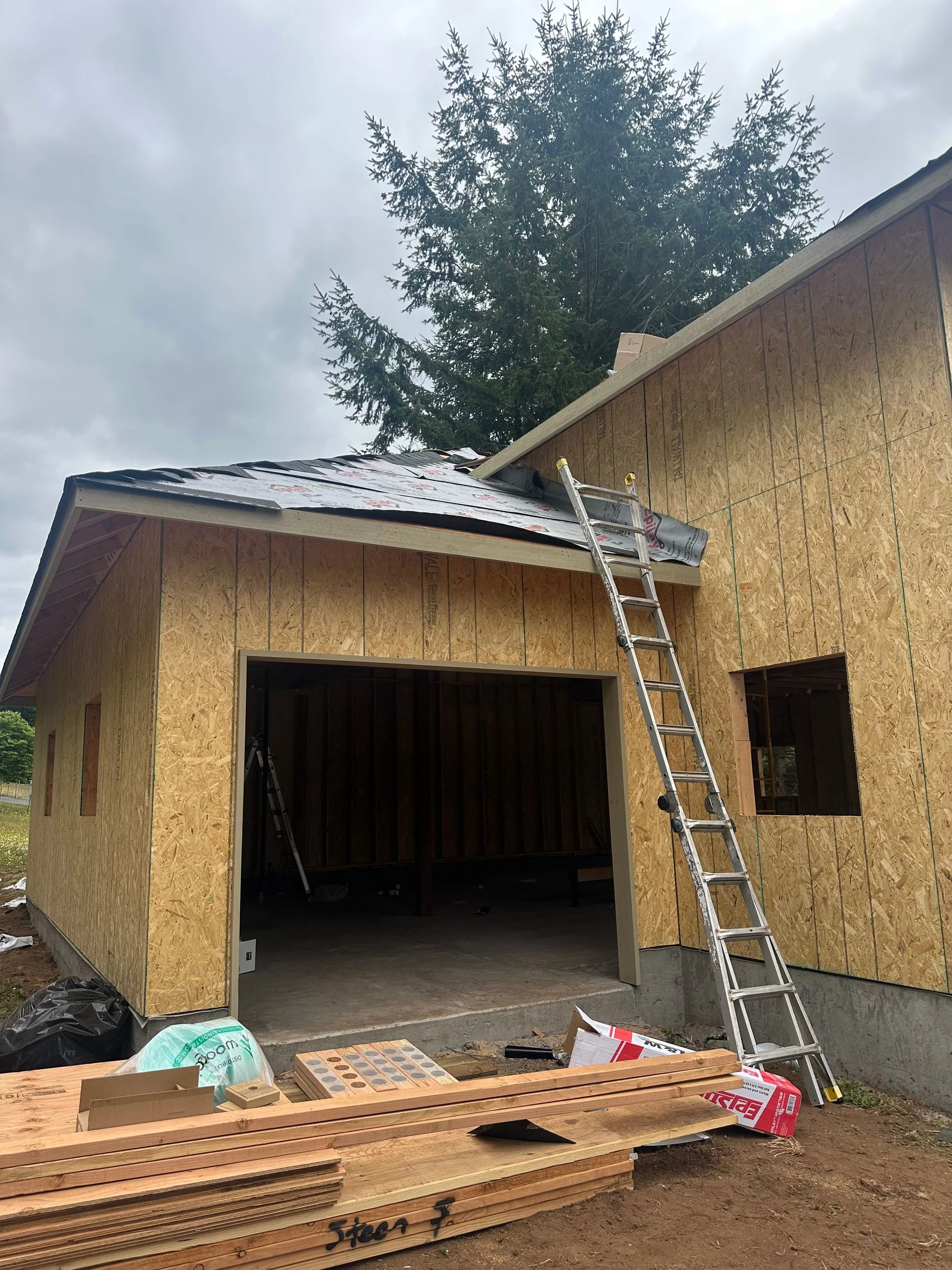 A garage under construction with plywood walls, a partially shingled roof, and a ladder leaning against the side.
