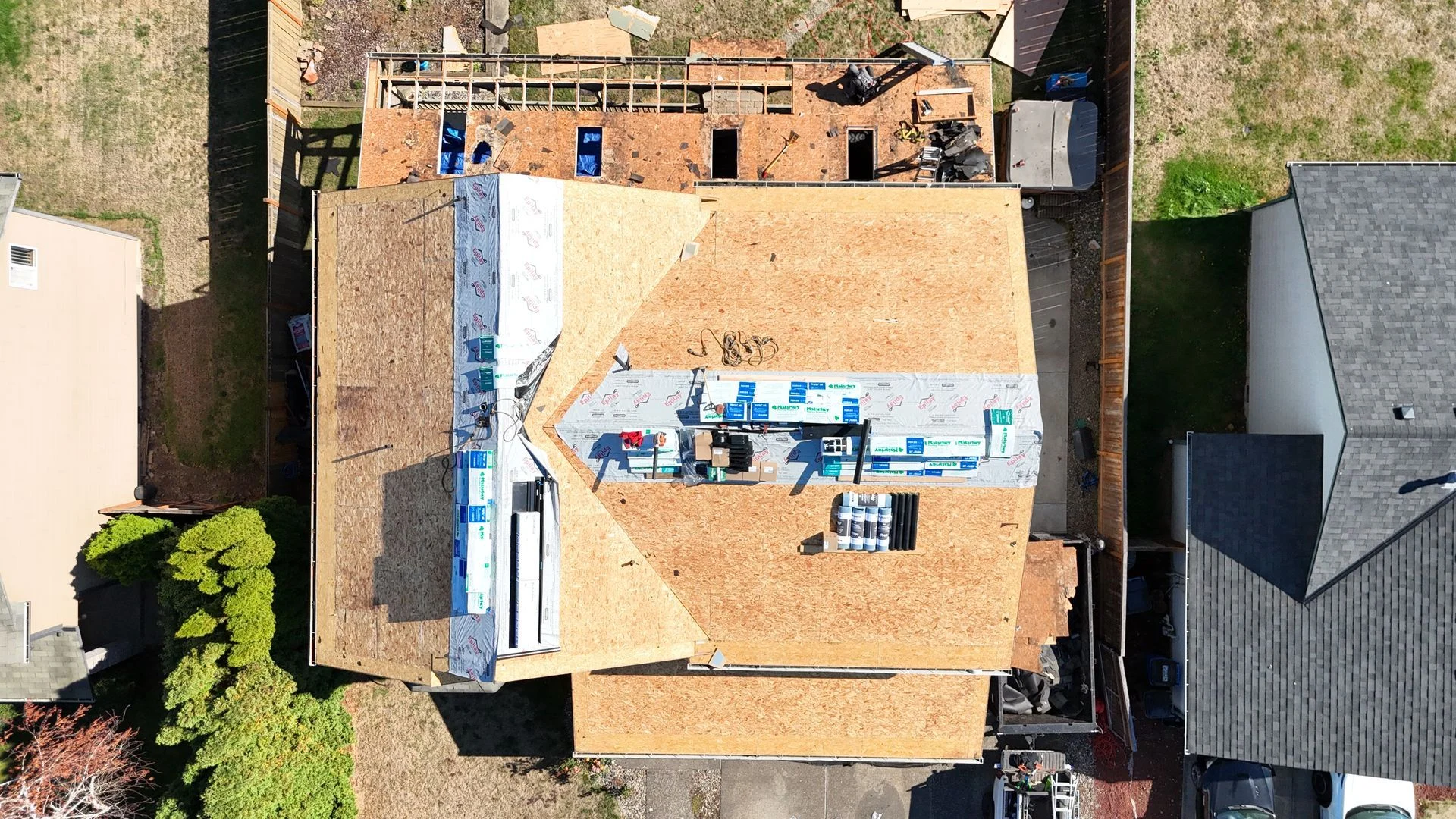 Aerial view of a house roof under construction, featuring plywood decking and sections of blue and grey underlayment.