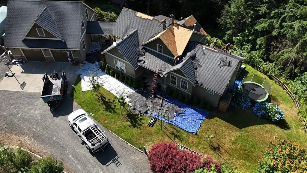 Roof replacement in progress, torn-off shingles on tarps, work van and dumpster in the drive