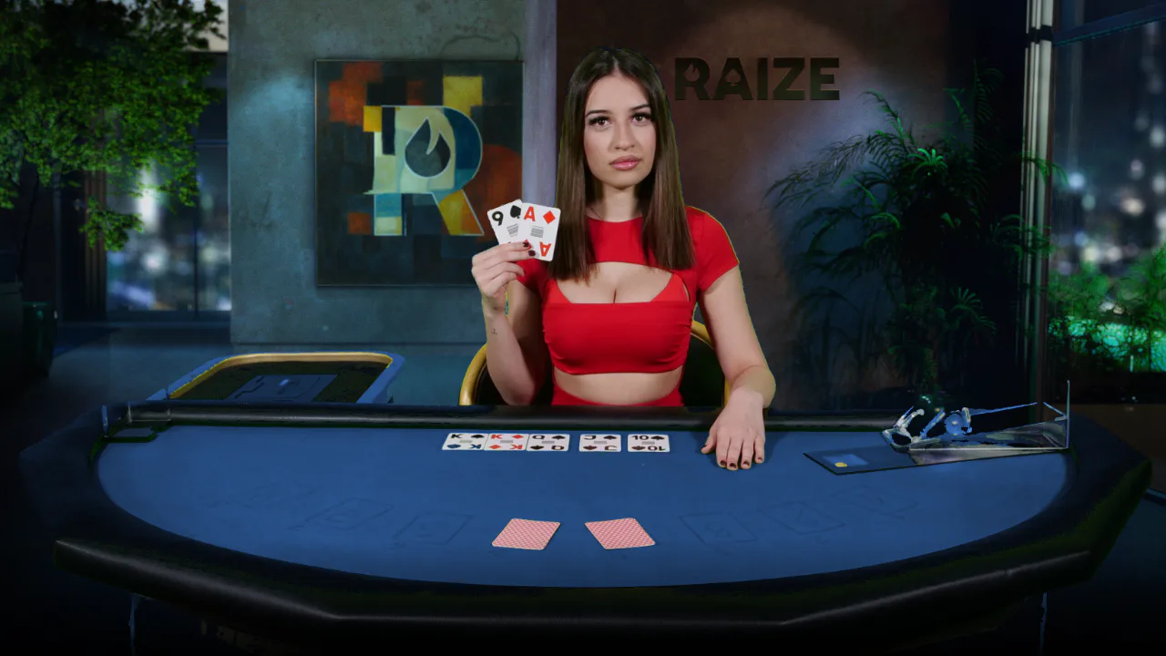 A RAIZE dealer in a red top holding up two cards at a blue felt table with five community cards dealt