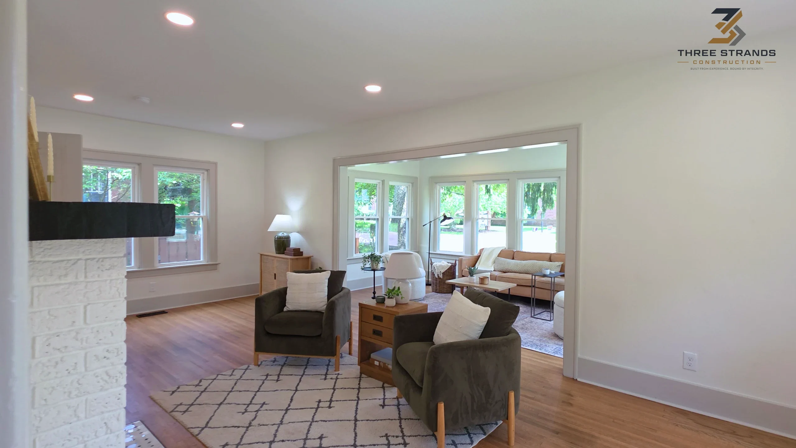 Renovated living room with white walls, hardwood floors, recessed lighting and a sunroom lined with windows, from a Three Strands Construction renovation video