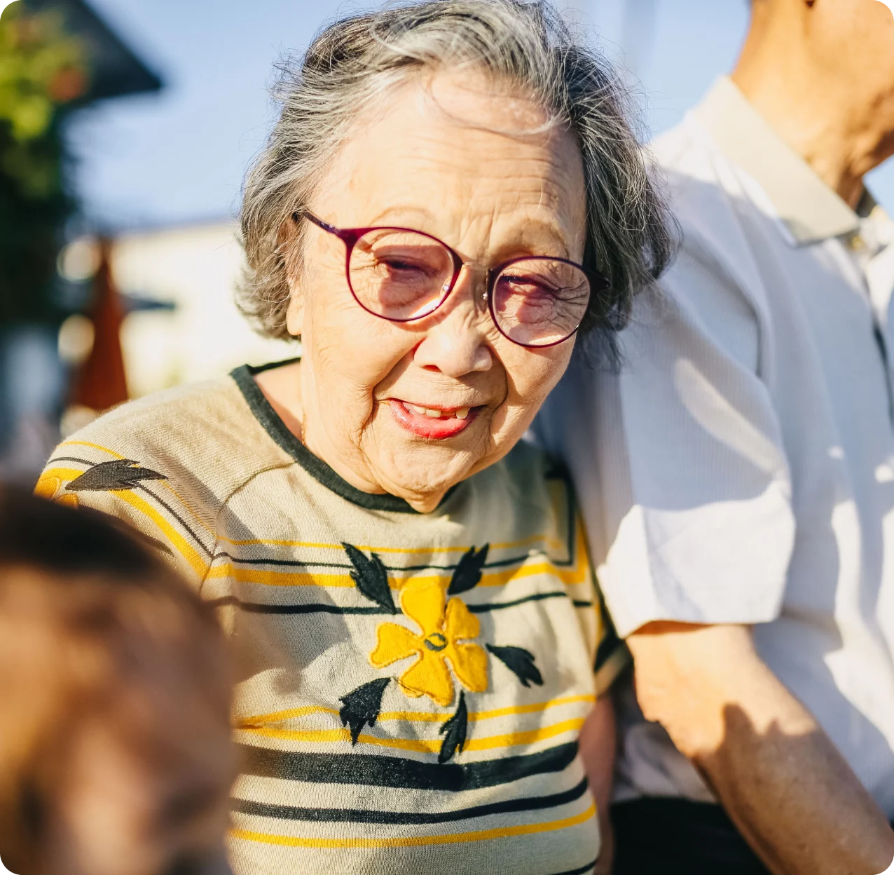An older woman smiling outdoors while spending time with her family.