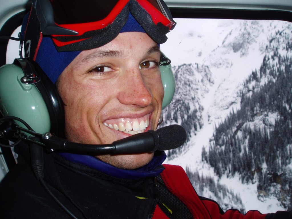 Kees in a helicopter, goggles up, grinning, with snowy peaks visible through the window.