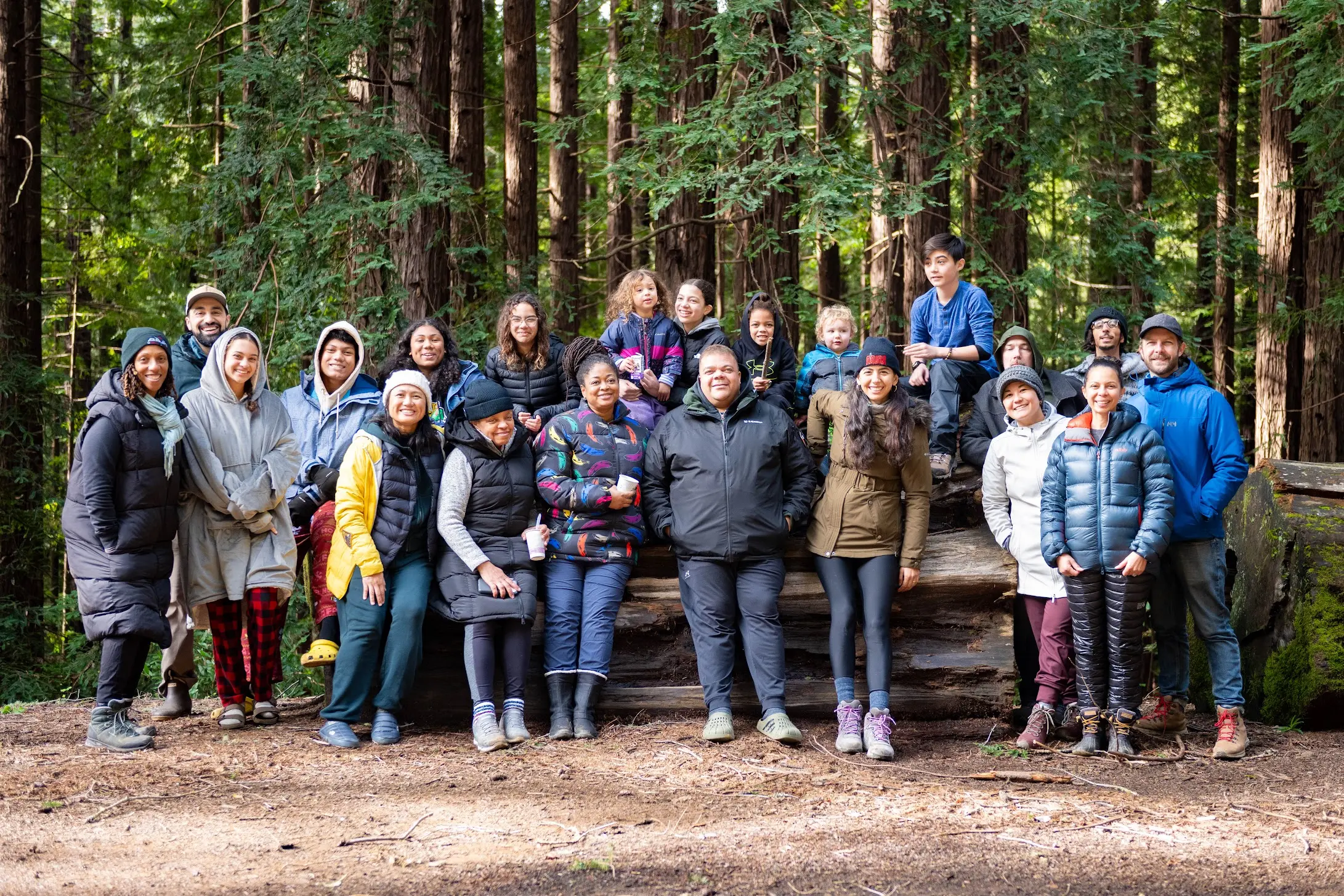 A multi-generational, multi-racial group of twenty-plus campers gathered on a fallen redwood in a California coastal forest — an Outdoorithm Collective trip.