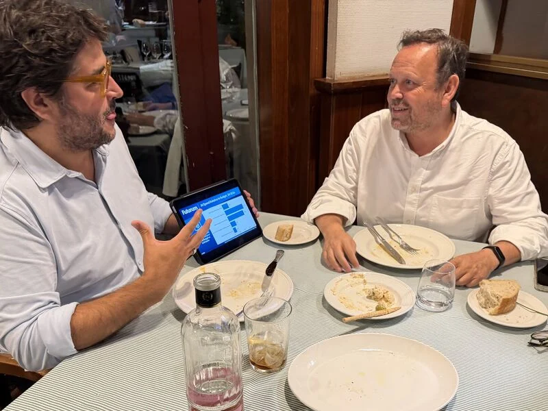 Rafael Tamames y Carlos Oliveira cenan en Madrid y comentan un gráfico sobre gasto en inteligencia artificial mostrado en un iPad.