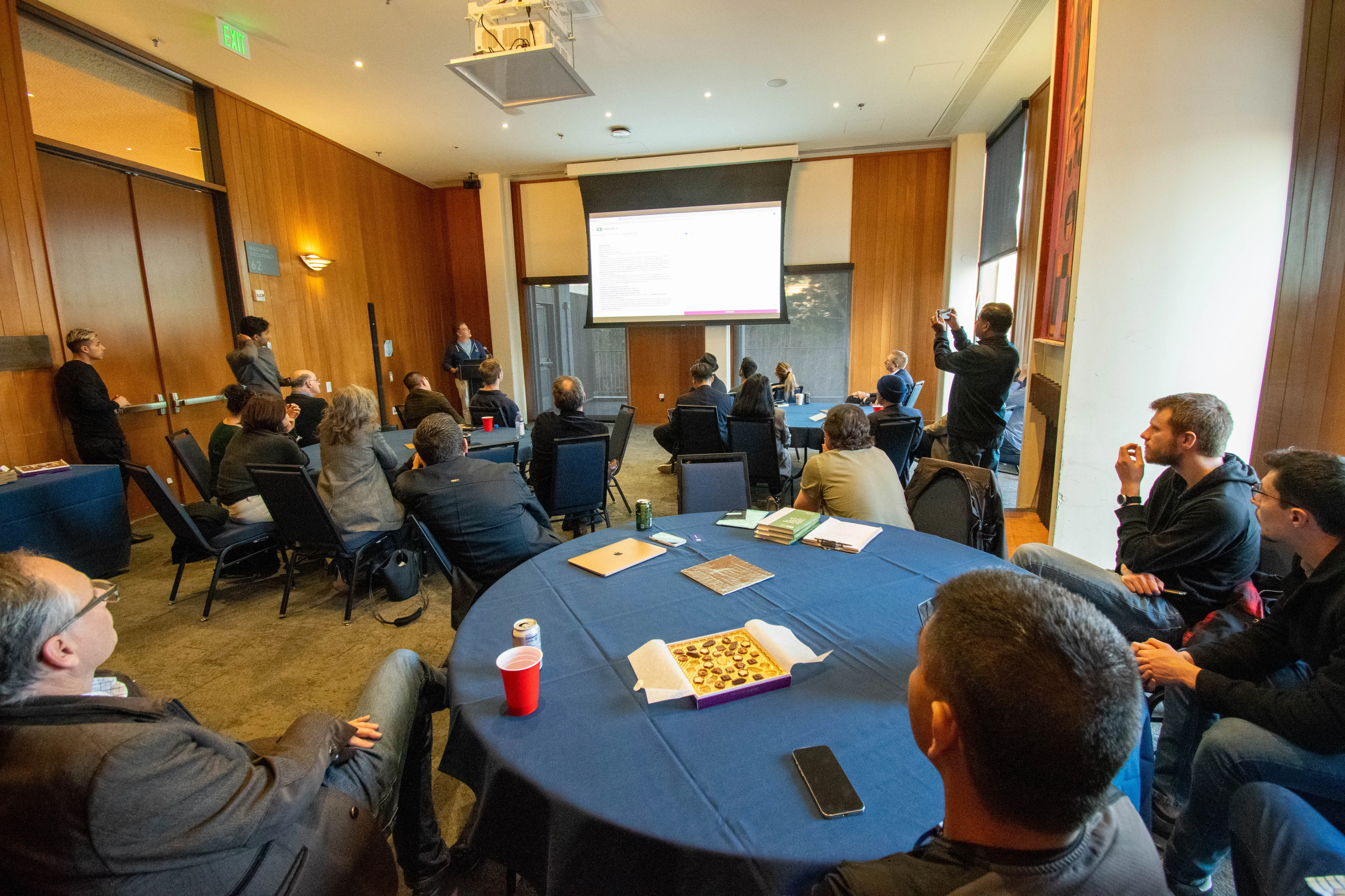 Audience seated at round tables facing a presenter and projection screen.