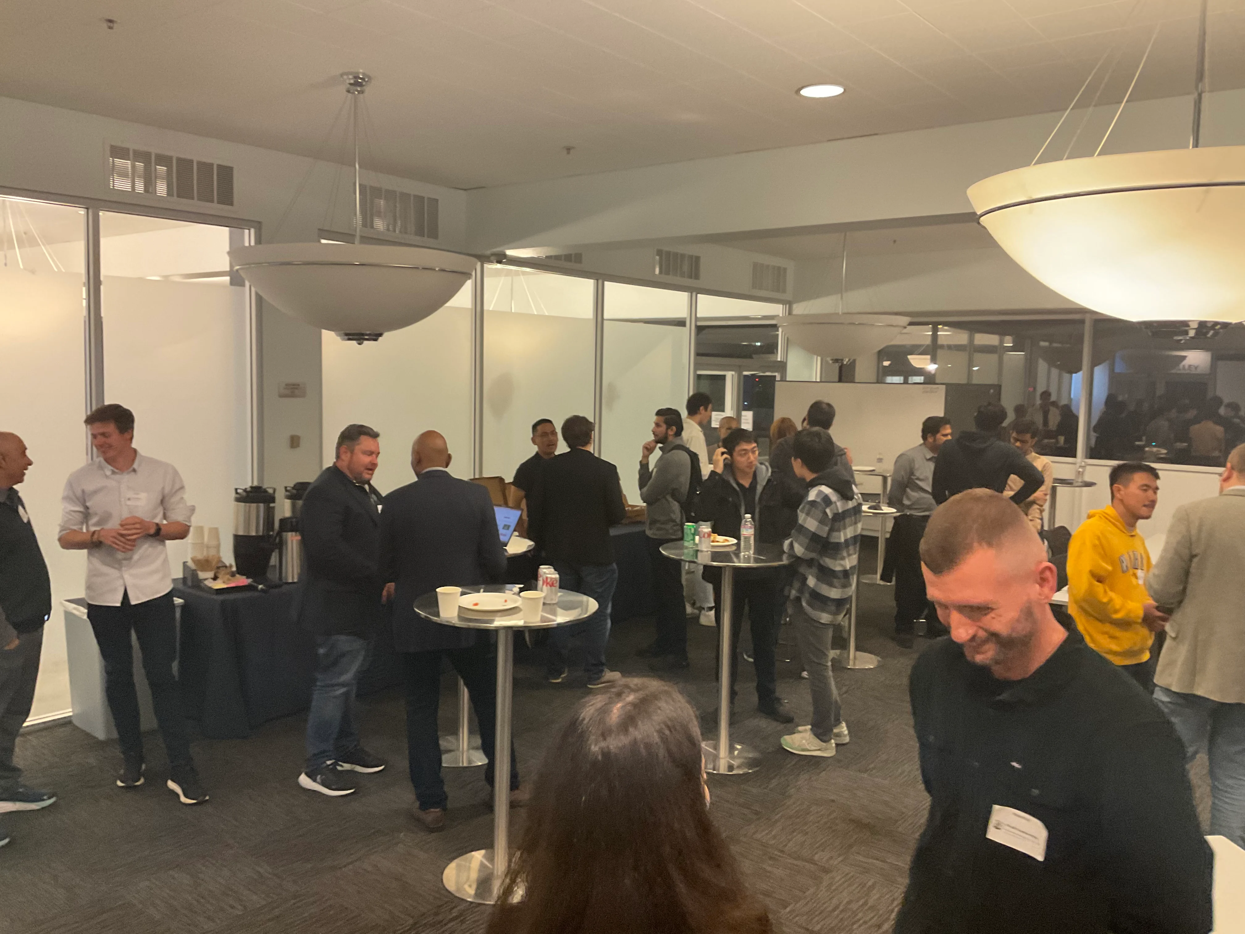 Attendees standing in conversation around high tables in a glass-walled room.