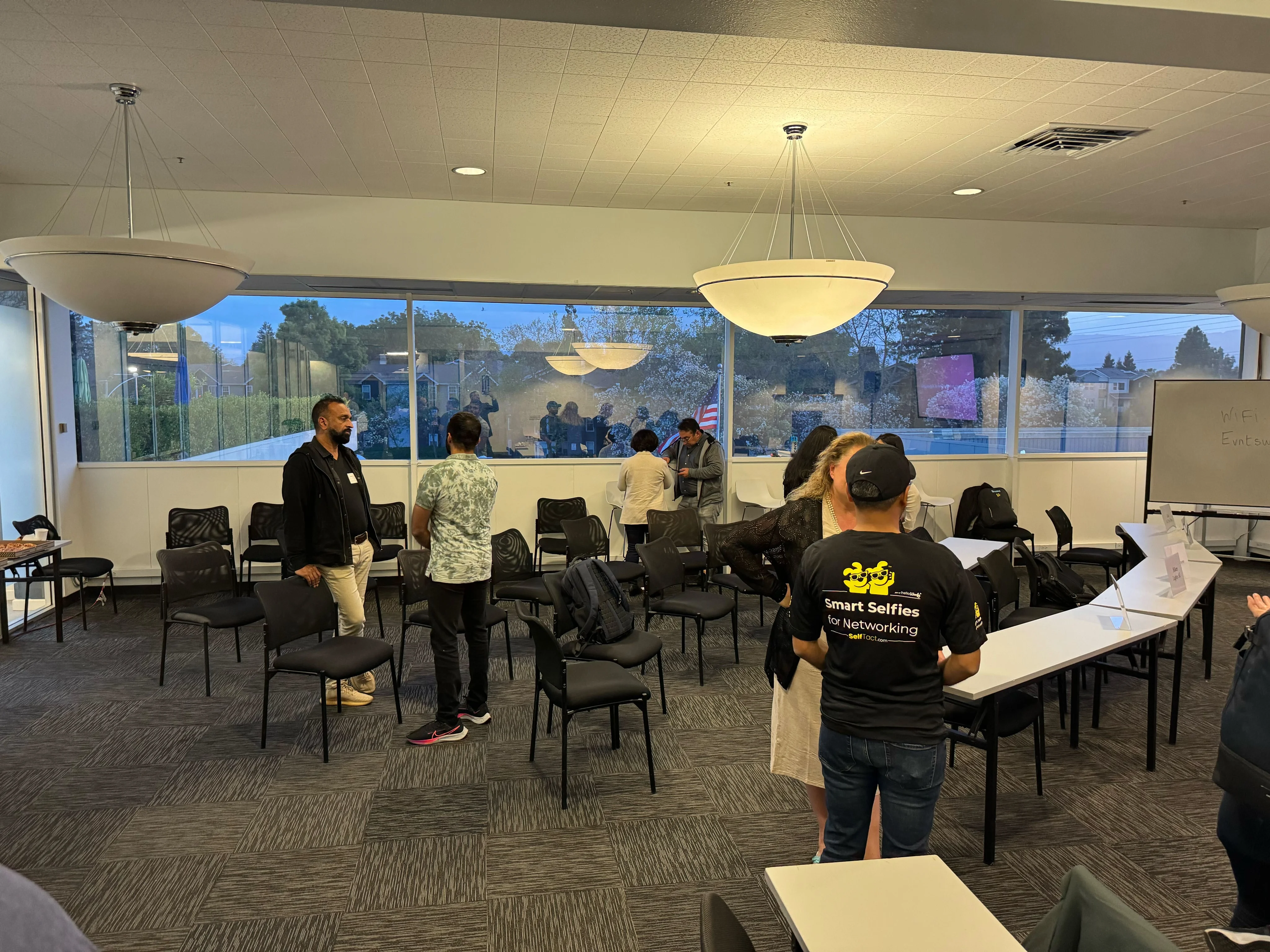 Attendees talking in small groups among chairs beside large windows.
