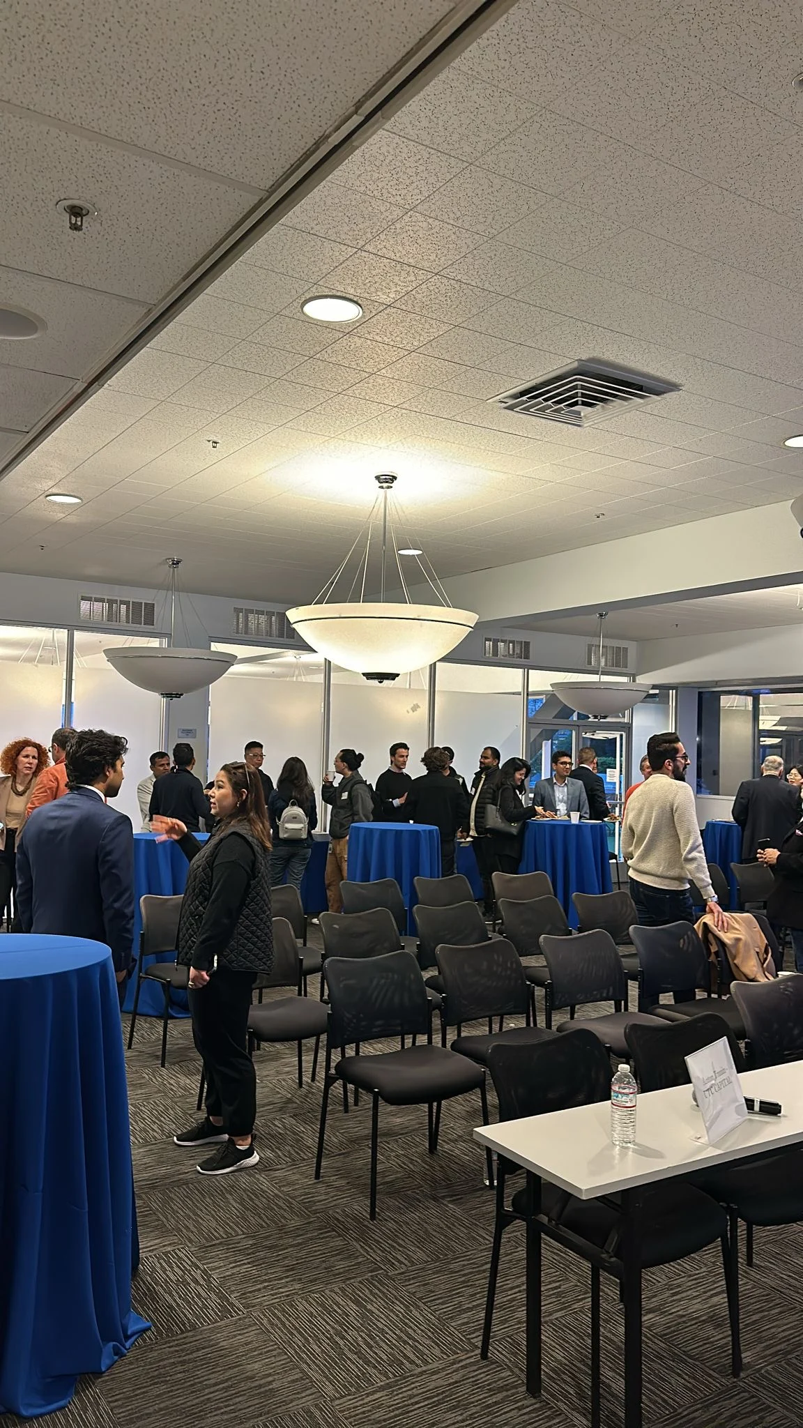 Attendees talking around blue-covered standing tables behind rows of chairs.