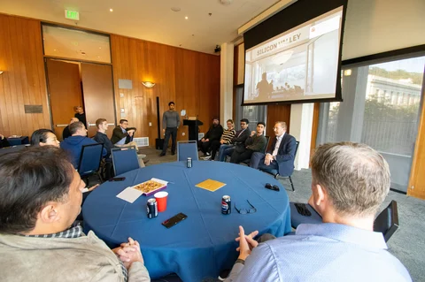 Participants seated around blue-covered tables during a discussion in a wood-panelled room.