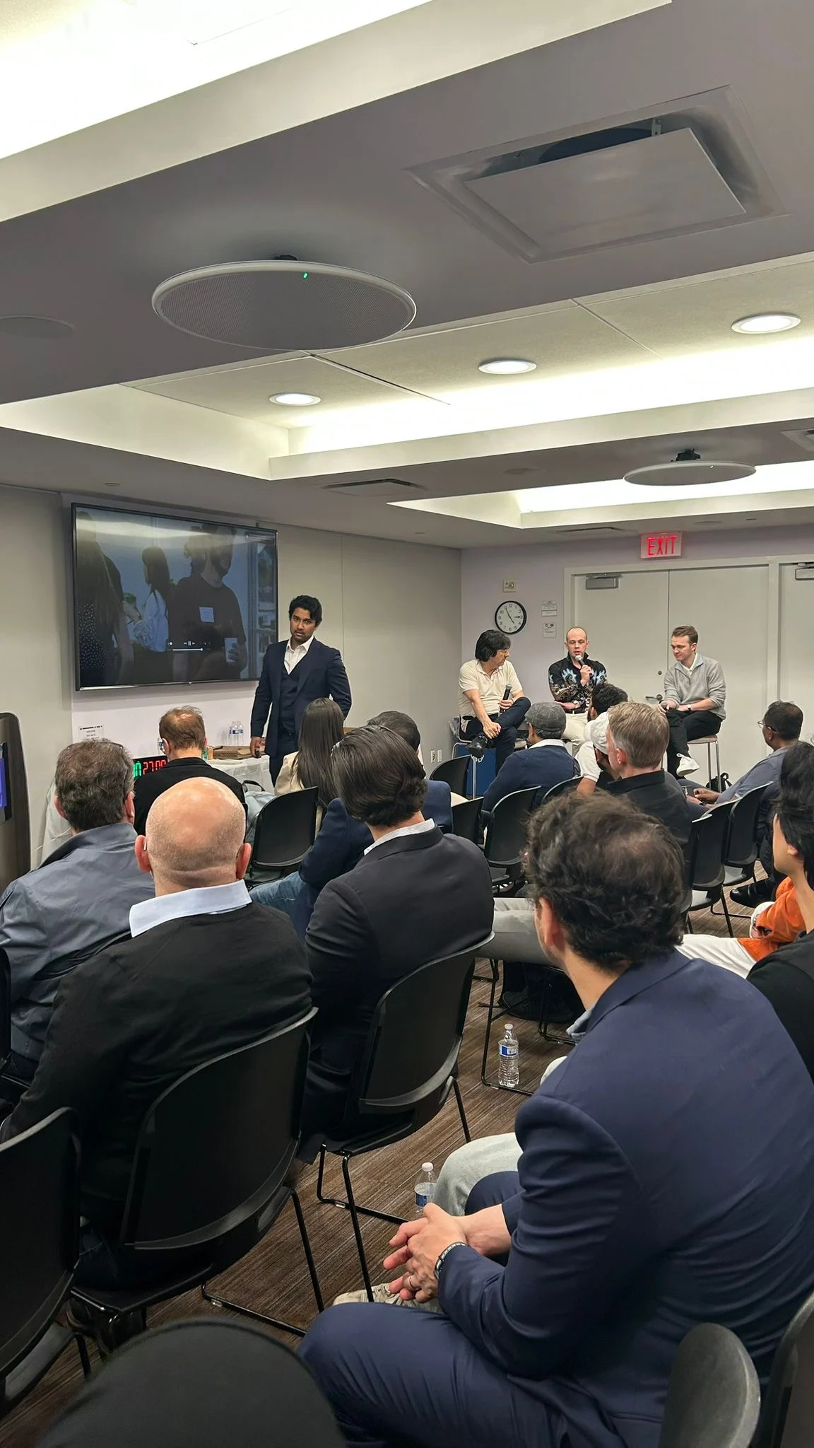 Seated audience facing a panel and a standing presenter in a meeting room.