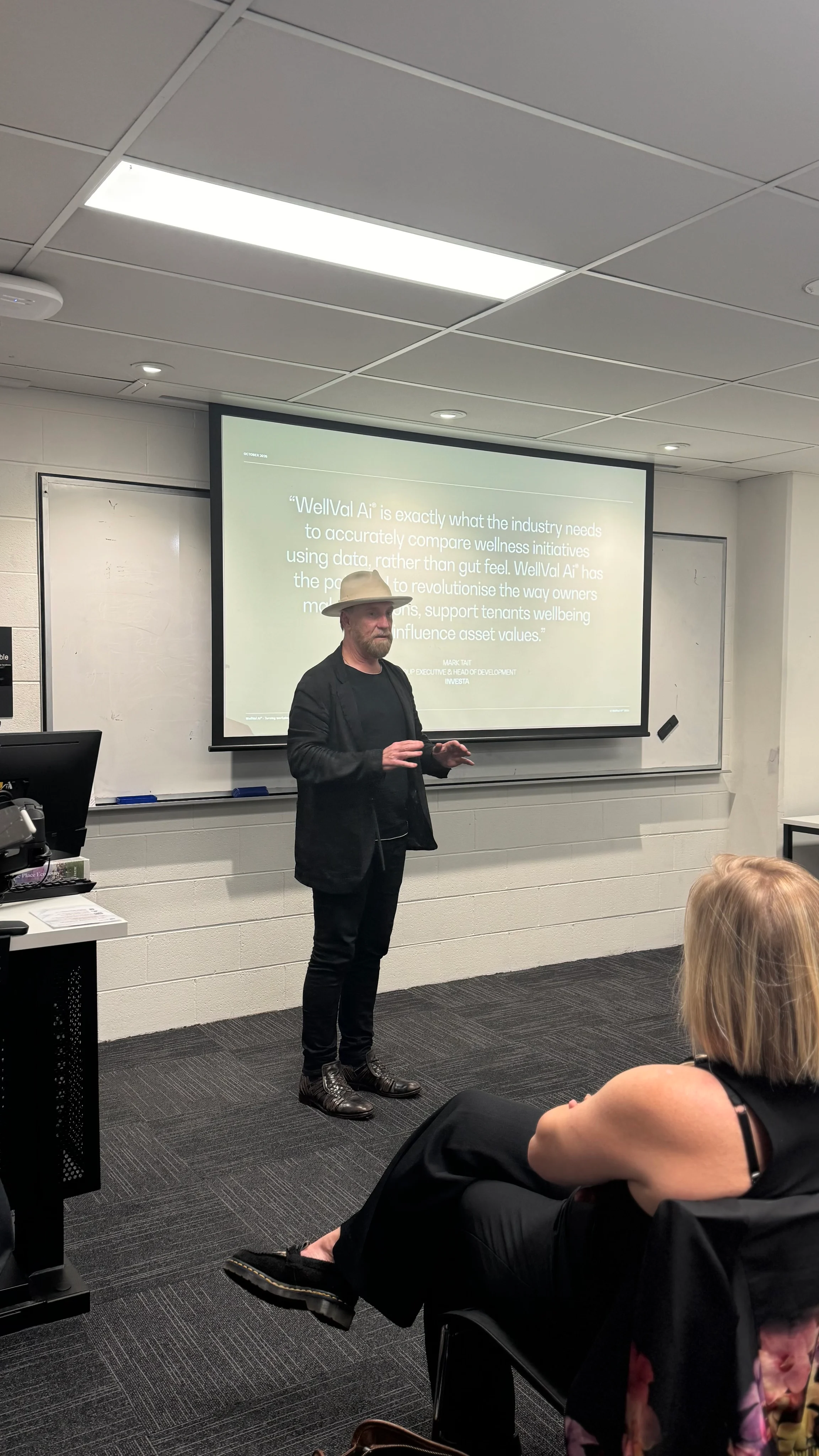 A speaker wearing a hat addressing attendees beside a projection screen.