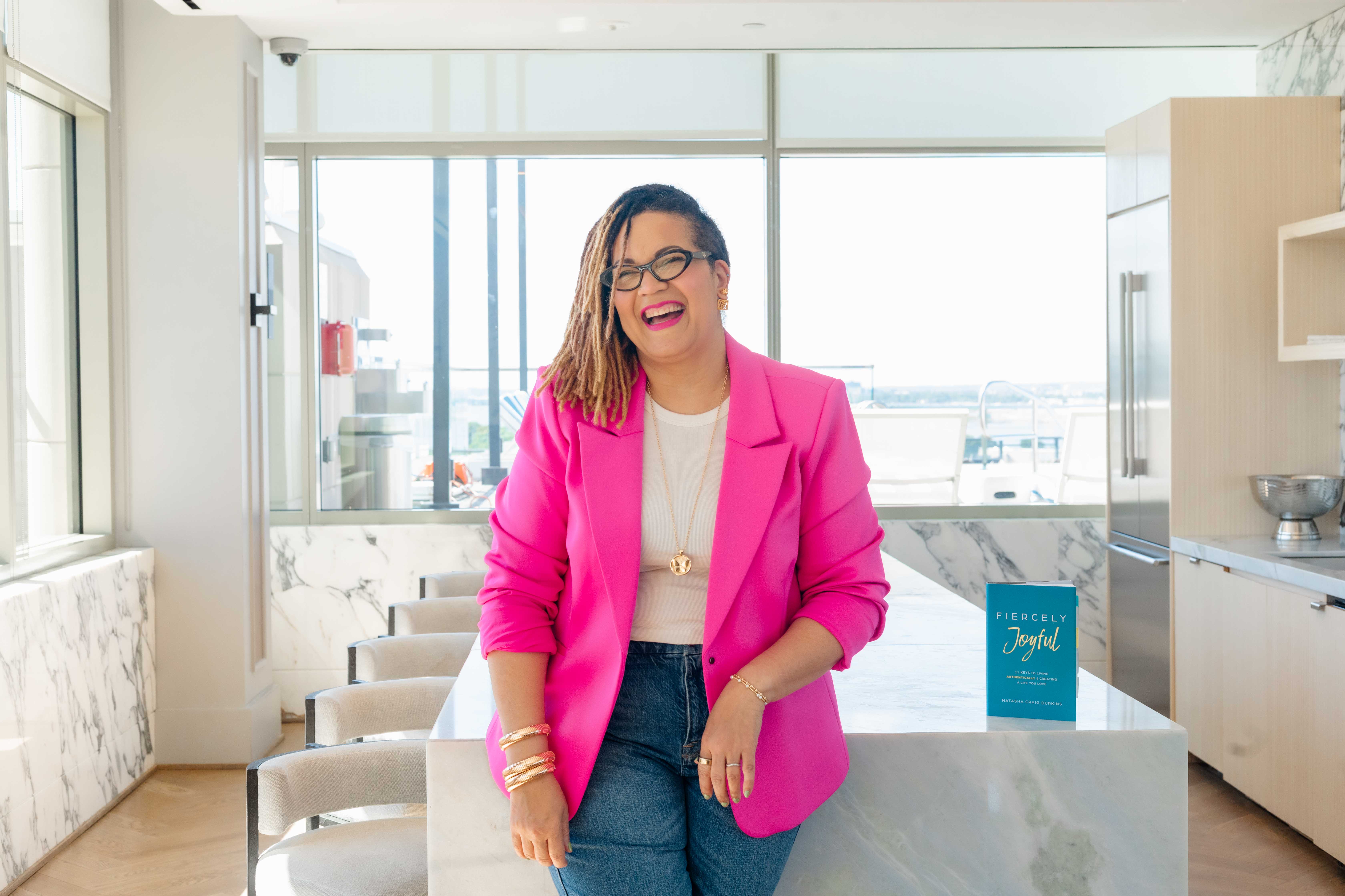 Tash Durkins laughing in a pink blazer beside her book Fiercely Joyful