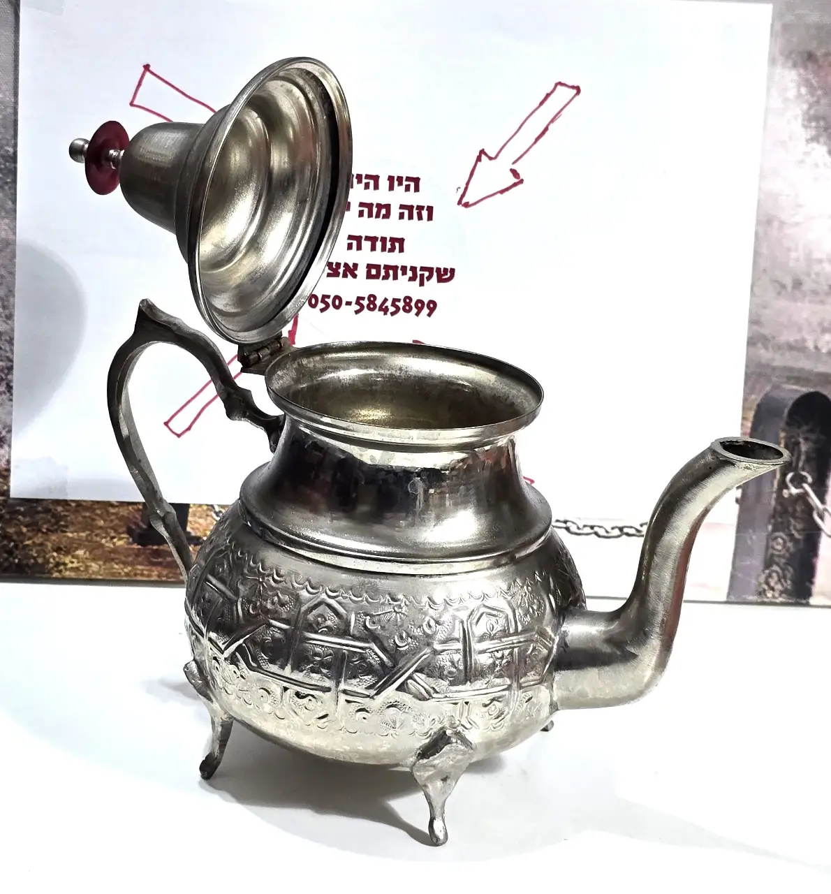 קומקום תה דקורטיבי בסגנון מזרחי ממתכת בגוון כסוף, עומד על שלוש רגליים, עם פיה ומכסה מחובר בציר. עיטור חרוט בתבליט בסגנון מסורתי. מתאים לאספנות, לעיצוב או לשימוש. מצב כללי טוב עם סימני שימוש בהתאם לגיל. מידות: ‎20×24×14 ס”מ