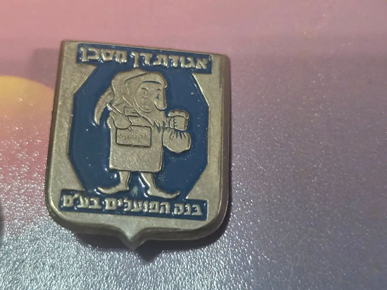 סיכה וינטג’ ממתכת מצופה אמייל, דמות מסורתית עם שק כסף, כיתוב “אגודתן חסכן” ו”בנק הפועלים בע״מ”. פריט פרסומי ישן בעיצוב אופייני לאמצע המאה ה-20. מצב טוב עם סיכה מקורית