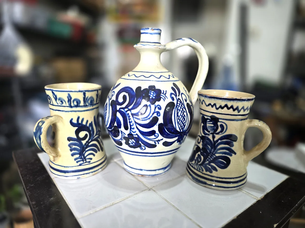 סט קרמיקה מסורתי הכולל קנקן (כד) ושני ספלים, המעוטרים במוטיבים פרחיים בכחול על רקע לבן.