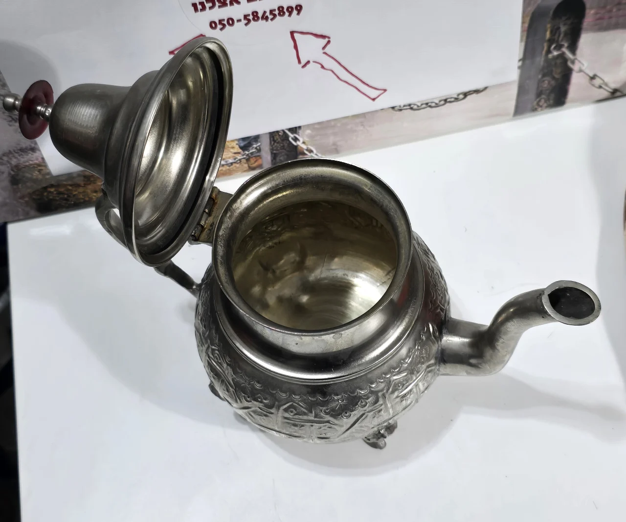 קומקום תה דקורטיבי בסגנון מזרחי ממתכת בגוון כסוף, עומד על שלוש רגליים, עם פיה ומכסה מחובר בציר. עיטור חרוט בתבליט בסגנון מסורתי. מתאים לאספנות, לעיצוב או לשימוש. מצב כללי טוב עם סימני שימוש בהתאם לגיל. מידות: ‎20×24×14 ס”מ