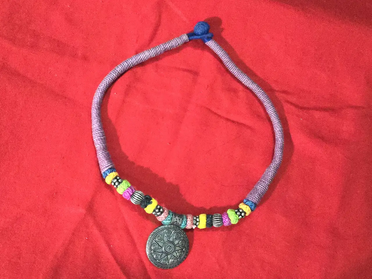 שרשרת בסגנון  אתני\שבטי השרשרת עשויה מחבל או חוט כותנה עבה עטוף בליפוף של חוטים צבעוניים משולבים בה חרוזי מתכת ותליון מתכת במרכז בעיצוב עתיק