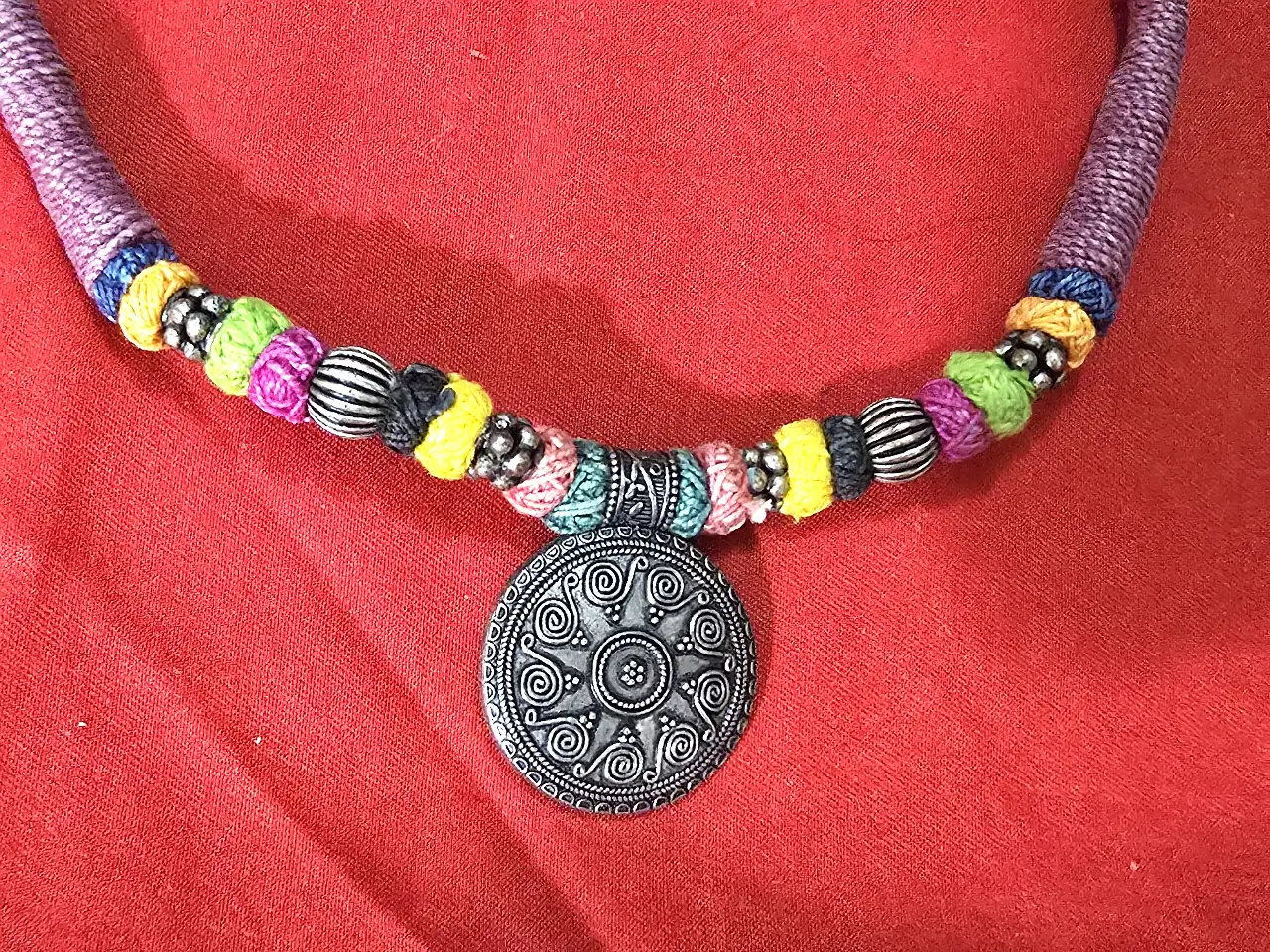 שרשרת בסגנון אתני\שבטי השרשרת עשויה מחבל או חוט כותנה עבה עטוף בליפוף של חוטים צבעוניים משולבים בה חרוזי מתכת ותליון מתכת במרכז בעיצוב עתיק