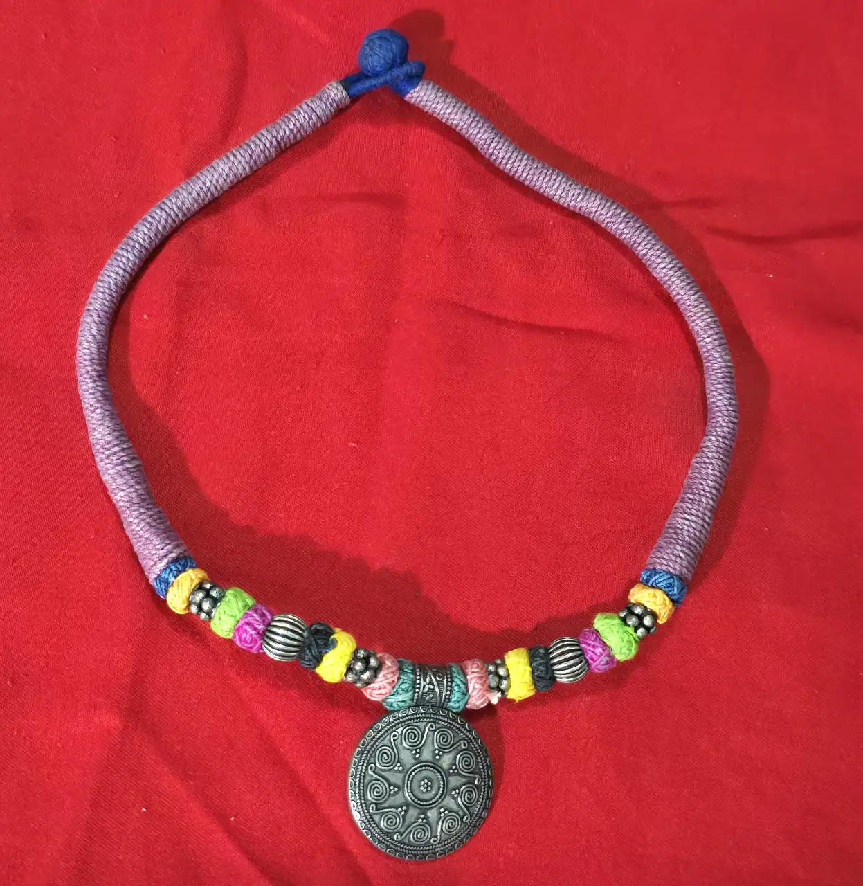 שרשרת בסגנון אתני\שבטי השרשרת עשויה מחבל או חוט כותנה עבה עטוף בליפוף של חוטים צבעוניים משולבים בה חרוזי מתכת ותליון מתכת במרכז בעיצוב עתיק