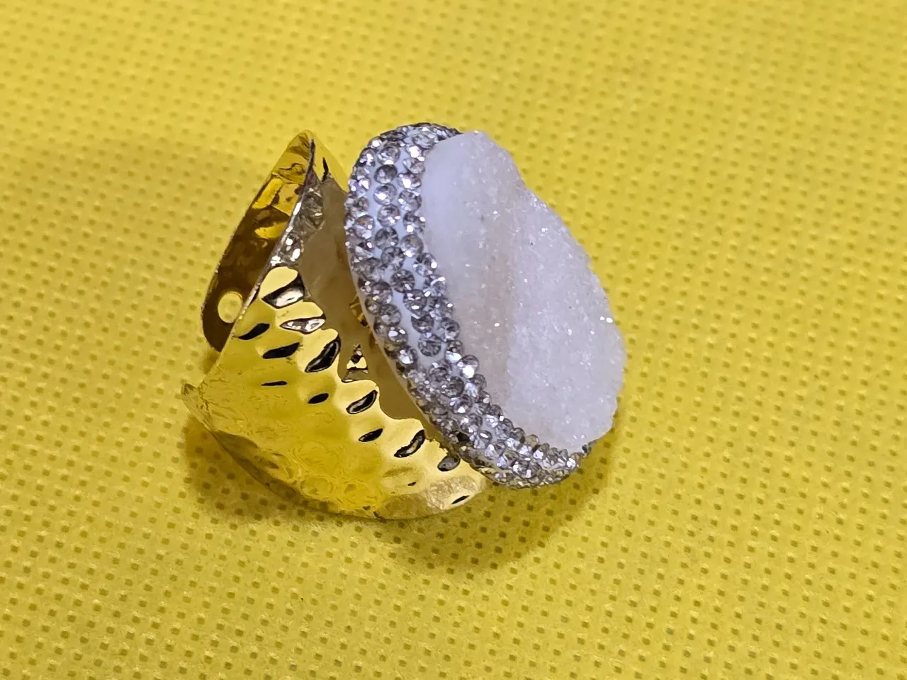טבעת מדהימה בעבודת יד! משובצת באבן מחוספסת גבישית נוצצת בצבעי לבן קרח. גוף מתכת מוזהב שניתן להתאים לגודל האצבע. מסביב יהלומים סינטטיים נוצצים.