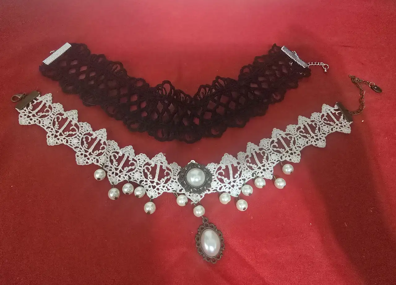 זוג שרשראות צ'וקר צמודות לצוואר שנהם מתחרה סגנון ויקטוריאני או גותי השרשרת הלבנה עשויה מתחרה לבנה בדוגמא גאומטרית שבחלק התחתון פנינים קטנות על גבי לולאות מתכת דקיקות התליון המרכזי משובץ בפנינה גדולה מעוטרת בהברונזה הצו'קר השחור מתחרה בדוגמת רשת עם סוגר ושרשרת הארכה כסוף