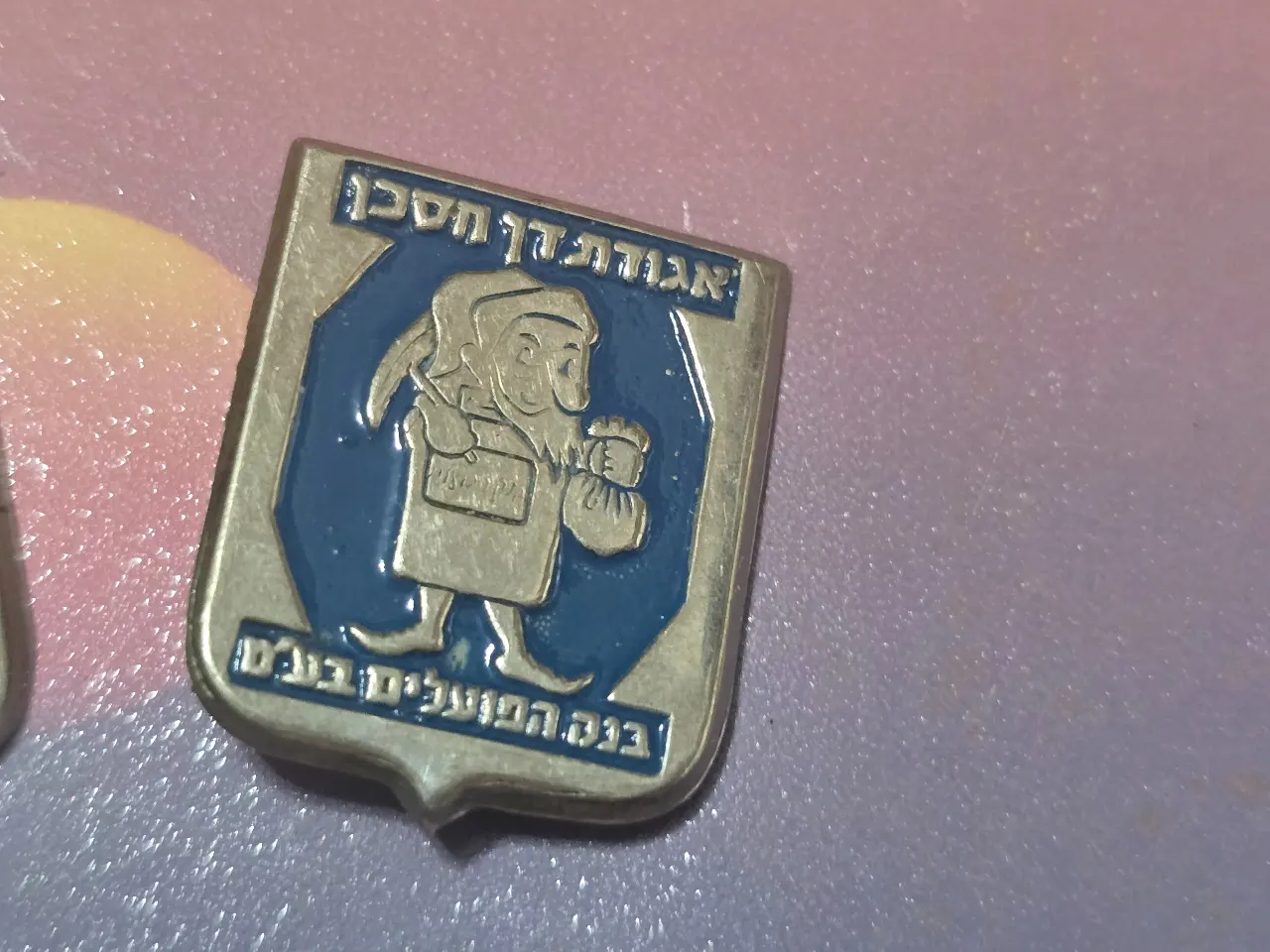 סיכה וינטג’ ממתכת מצופה אמייל, דמות מסורתית עם שק כסף, כיתוב “אגודתן חסכן” ו”בנק הפועלים בע״מ”. פריט פרסומי ישן בעיצוב אופייני לאמצע המאה ה-20. מצב טוב עם סיכה מקורית