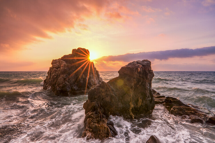 Umjetnička fotografija Rays of sunlight at sunset with rocks in rough sea