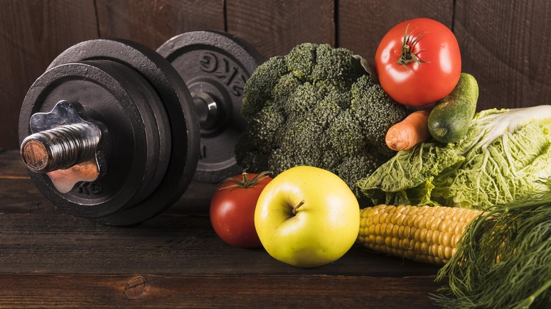 barra de exercícios ao lado de tomate, maçã, brócolis, milho, cenoura, pepino e couve sob a mesa