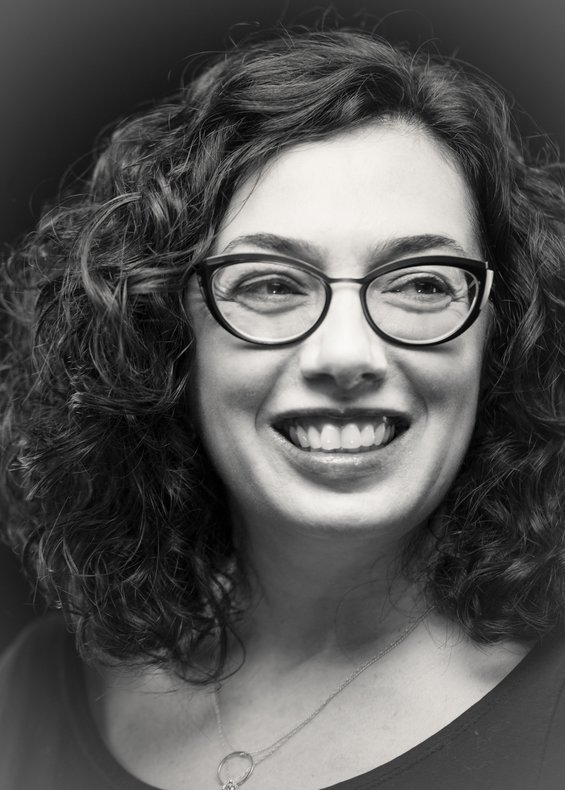 Helen Maria is shown in black & white looking to the right smiling wearing glasses with dark, curly hair. She is wearing a dark scoop-neck t-shirt with a small, thin necklace with a circle charm.