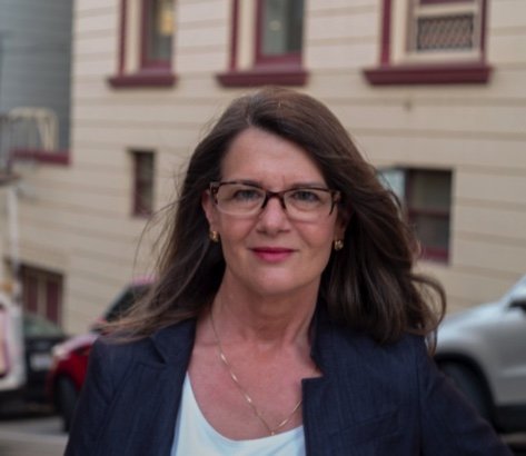 Suzanne is in a navy blue blazer with a white shirt, red lipstick, glasses, and long brown hair in front of a yellow building with red windows.