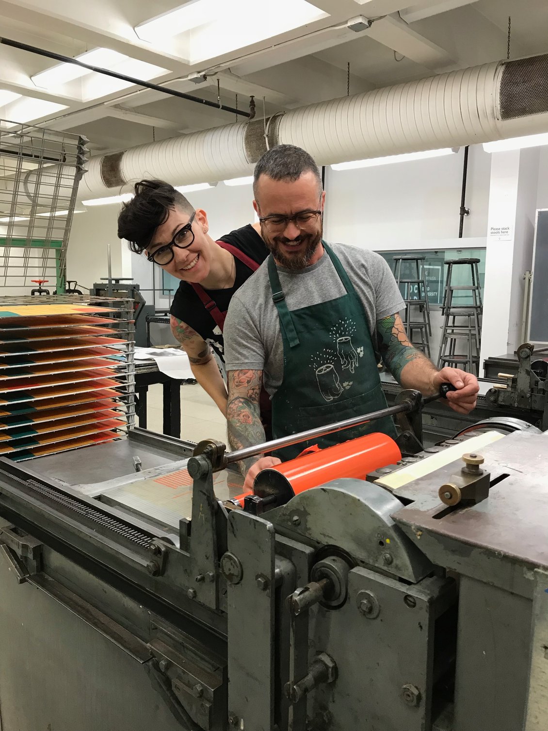 Printing press with Professor Anthea Black, who has short black hair wearing glasses, a black shirt and red apron, working with another individual wearing glasses in a grey shirt and blue apron, to roll a piece through.