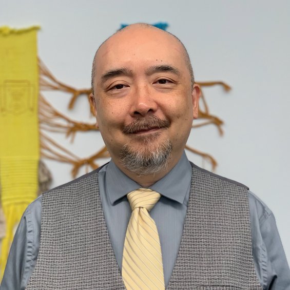 Maxwell in a soft yellow tie, light grey vest, and light grey-blue button down shirt, smiling in front of an art piece.