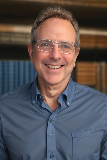 Man with silver glasses wearing blue button up shirt poses for headshot photo with library backdrop