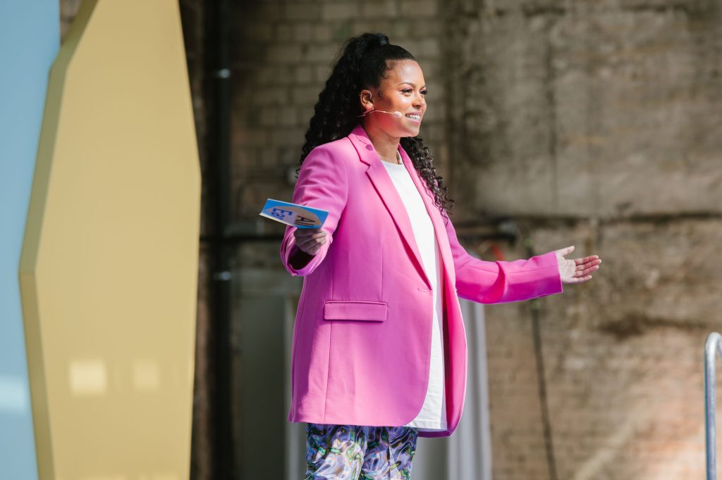 Moderator Aminata Belli is standing on the Spotify All Ears Summit main stage with her arms open wide as she talks to the audience