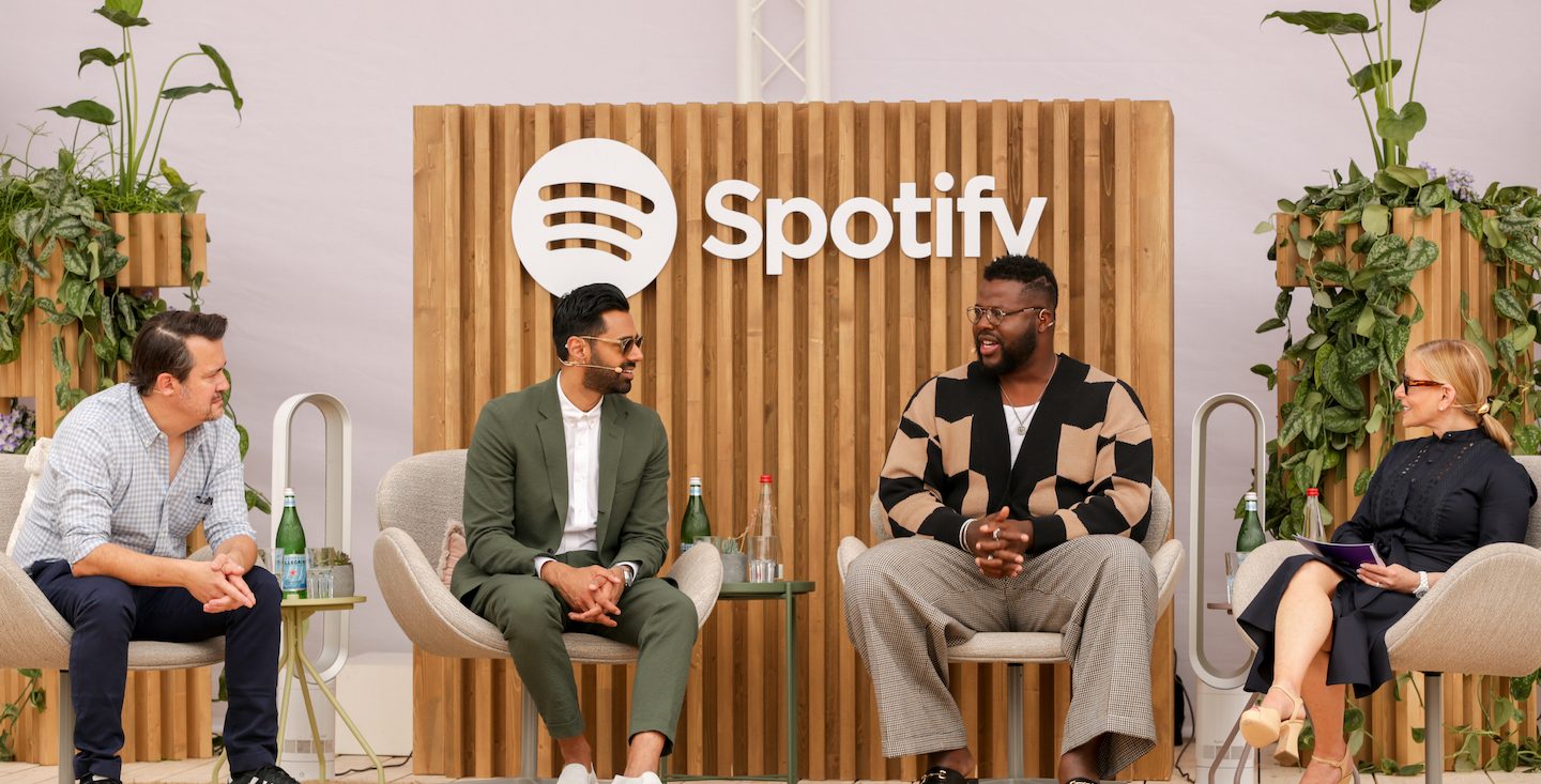 Peter Girardi, Hasan Minhaj, Winston Duke and Dawn Ostroff sit outside on a stage in front of a spotify logo. they are having a conversation.