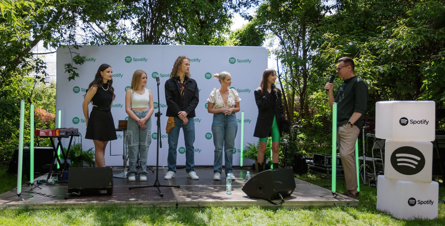 five singers standing on stage outside listening to a person speak