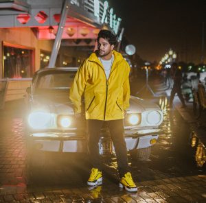 Asim Azhar standing in front of a car with its headlights on at night.