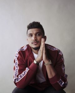 DIVINE looking straight at the camera with his hands in a prayer gesture