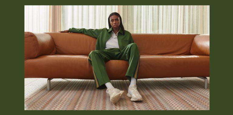 a photo of Cat Burns sitting on a brown leather couch, wearing a green tracksuit, and staring at the camera