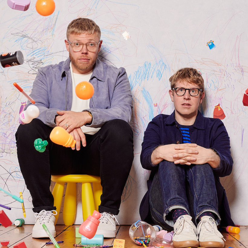 a photo of the parenting hell hosts sitting against a wall with toys thrown about